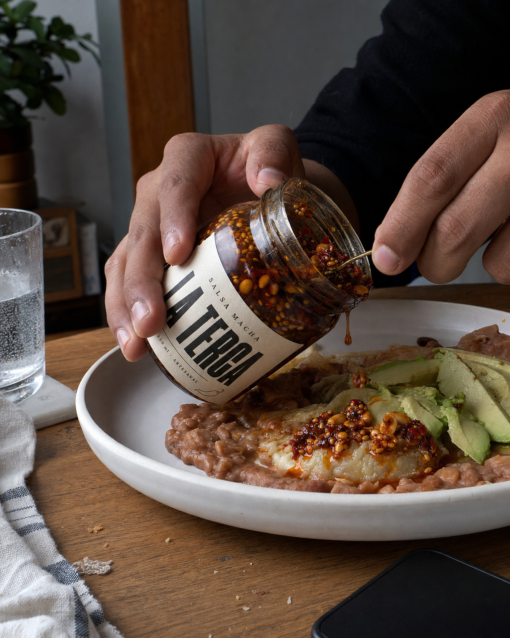 Unas manos vuelcan el frasco sobre un plato de comida en una mesa de casa, con luz de ventana
